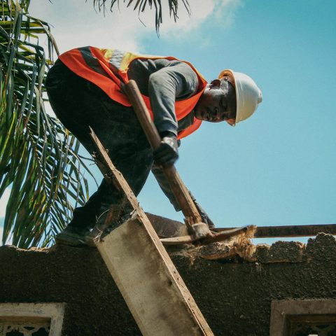 Construction worker demolishes roof under bright sky. Safety gear visible.