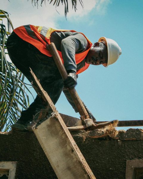 Construction worker demolishes roof under bright sky. Safety gear visible.