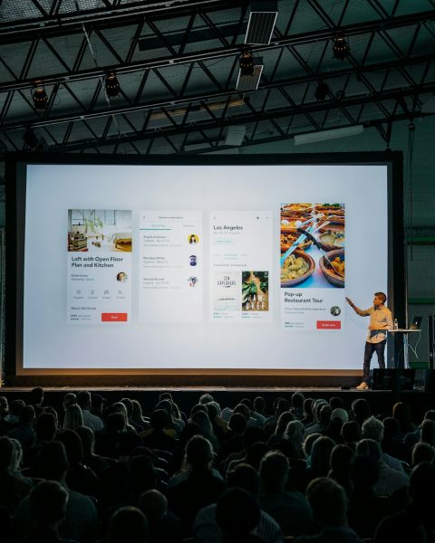 person discussing while standing in front of a large screen in front of people inside dim-lighted room